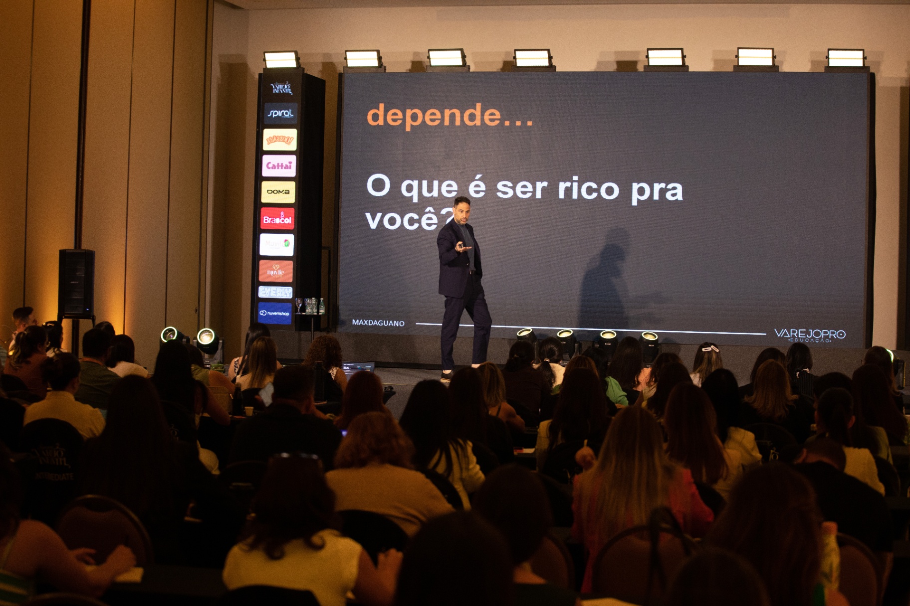 Max Daguano apresentando em palco com telão para plateia de varejo